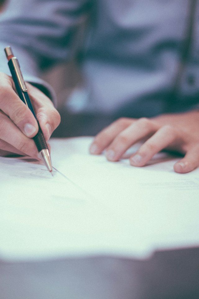 Mão segurando uma caneta sobre documentos impressos, em uma mesa de trabalho